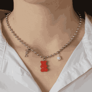 Collier argenté à billes porté autour du cou, orné de trois charms pendentifs : une clé, un ourson rouge et une perle, style délicat et romantique